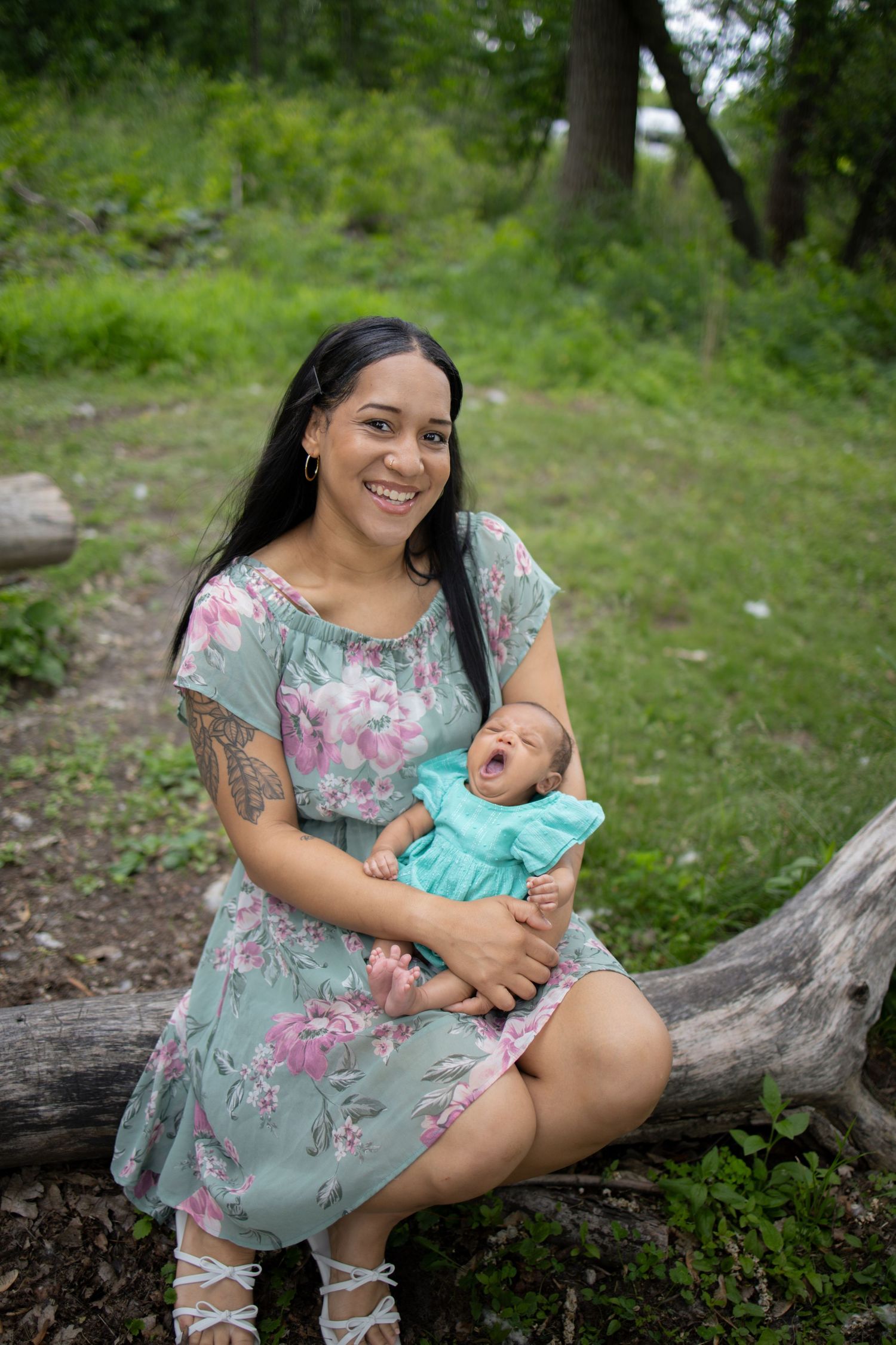A woman in a floral dress sits on a fallen log in a green outdoor setting while holding a baby in turquoise clothing.