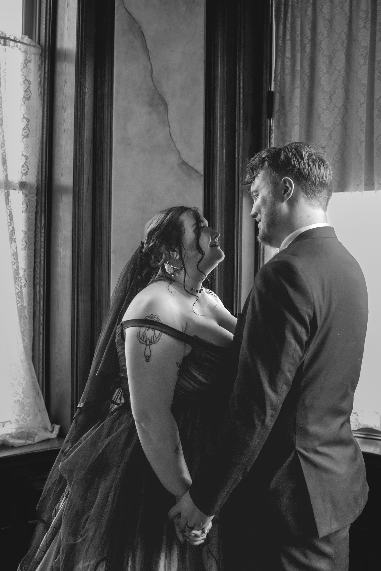 A romantic black and white photo of a couple in formal attire sharing an intimate moment by a window.