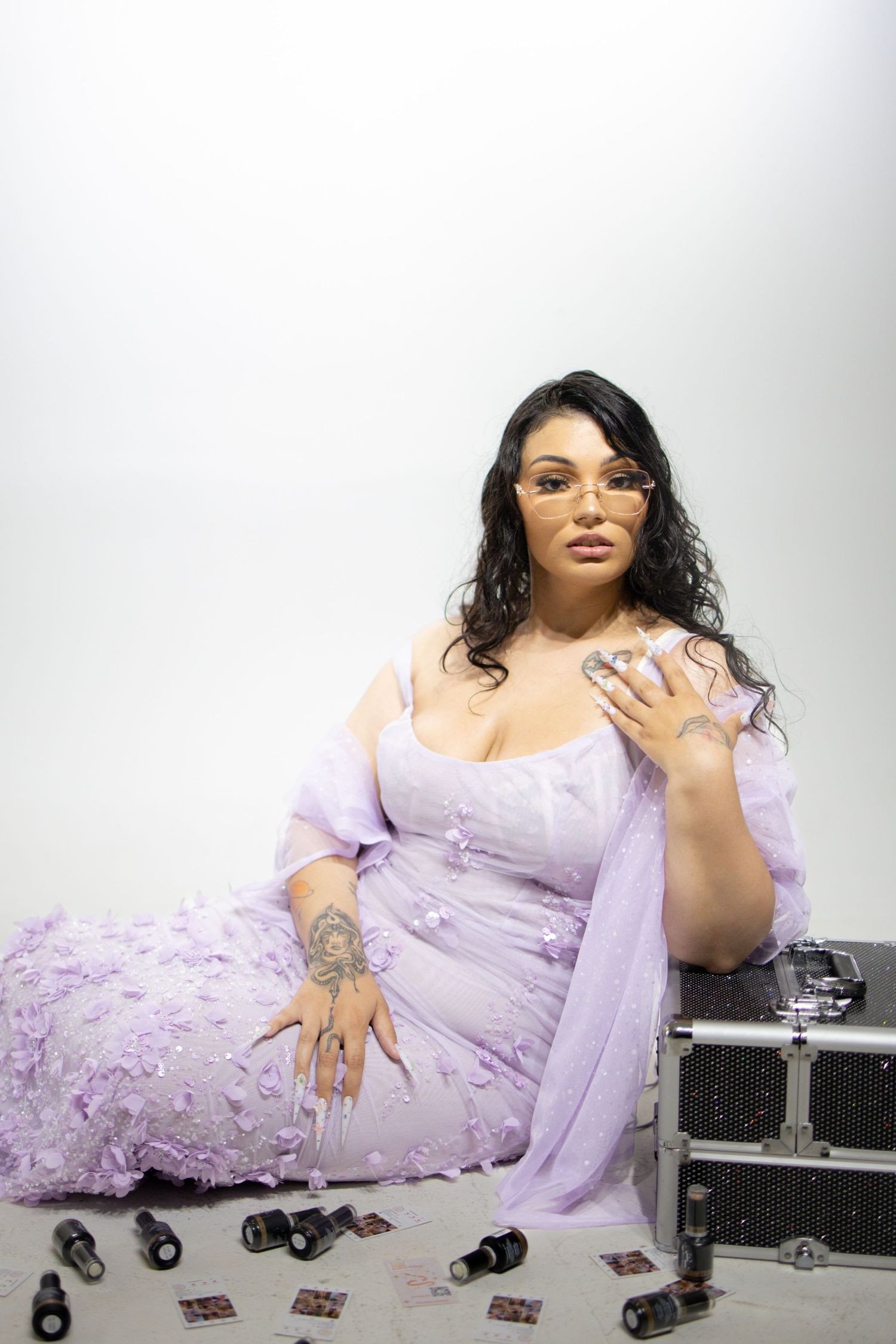 A fashion model in a flowing lavender dress poses elegantly on the floor against a white backdrop with black cases.