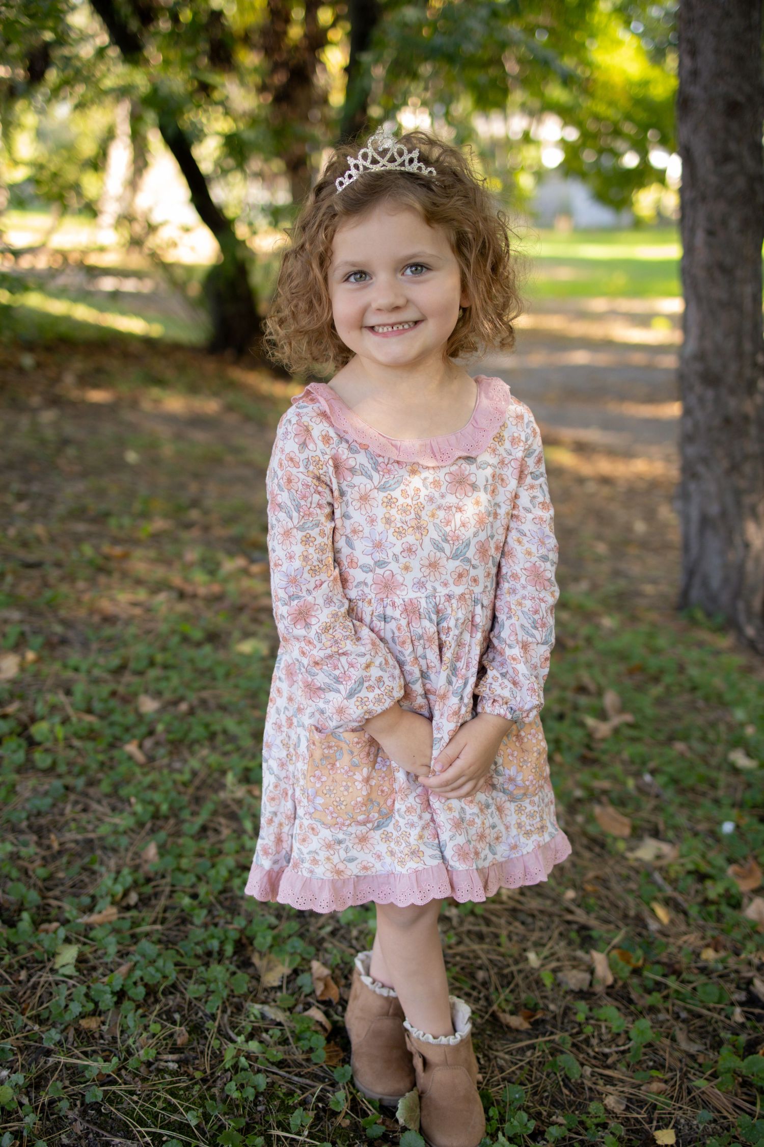 A young child wearing a pink floral dress and tiara poses outdoors in a park setting during autumn.