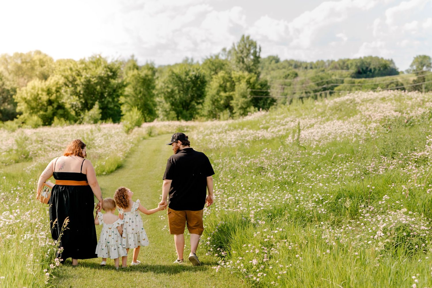 Three people walking hand in hand along a wildflower-filled meadow trail on a sunny summer day.