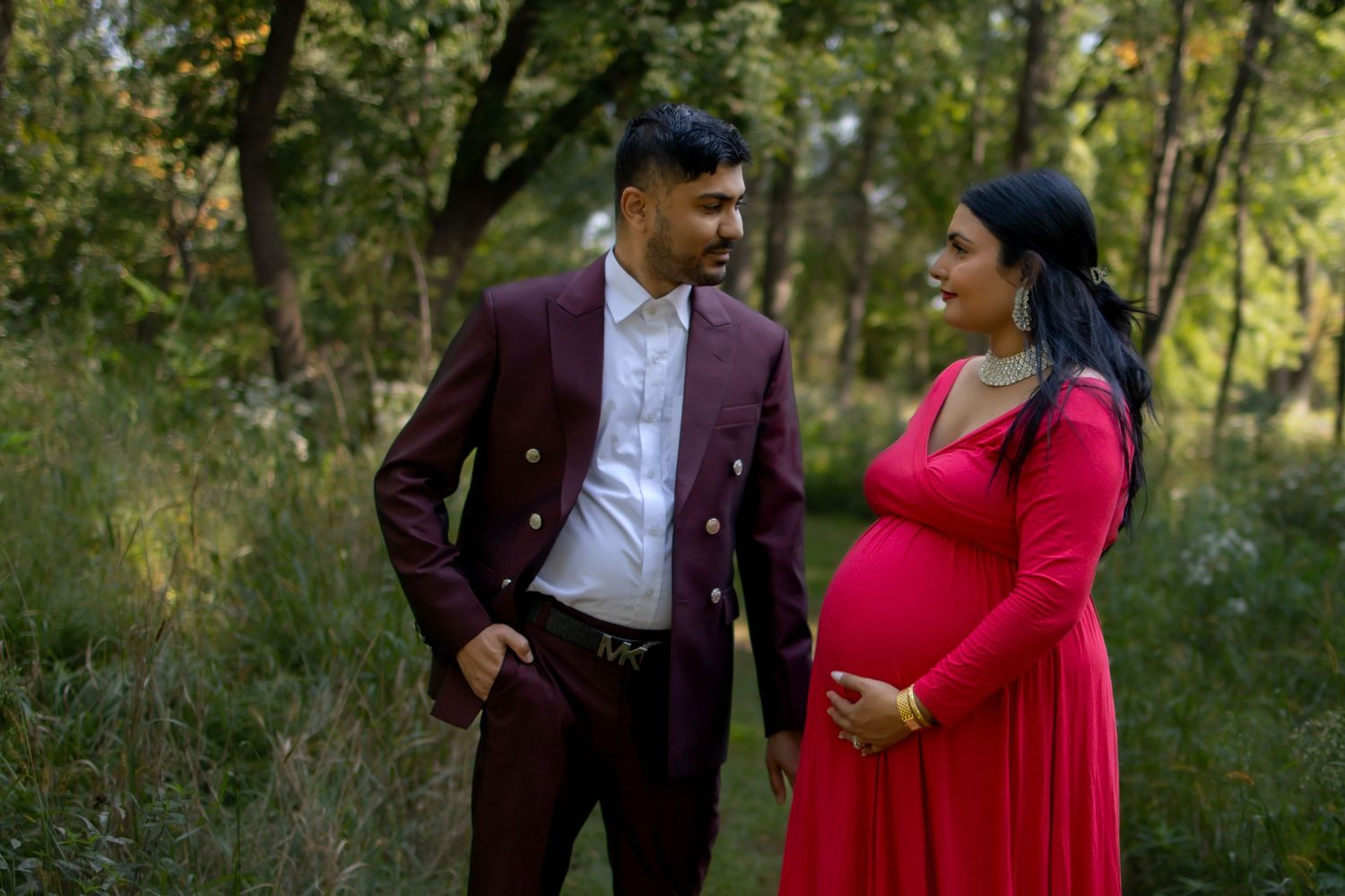 A couple walks together through a lush green outdoor setting, the woman wearing a vibrant red dress.
