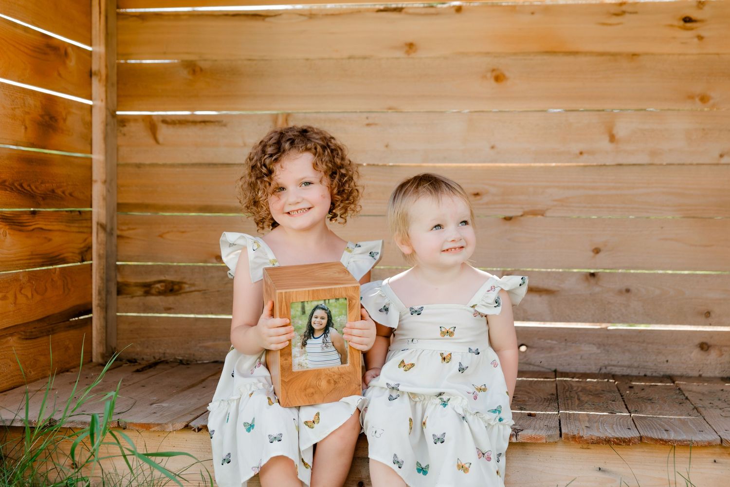 Two young children in floral outfits sit against a wooden wall holding photos.