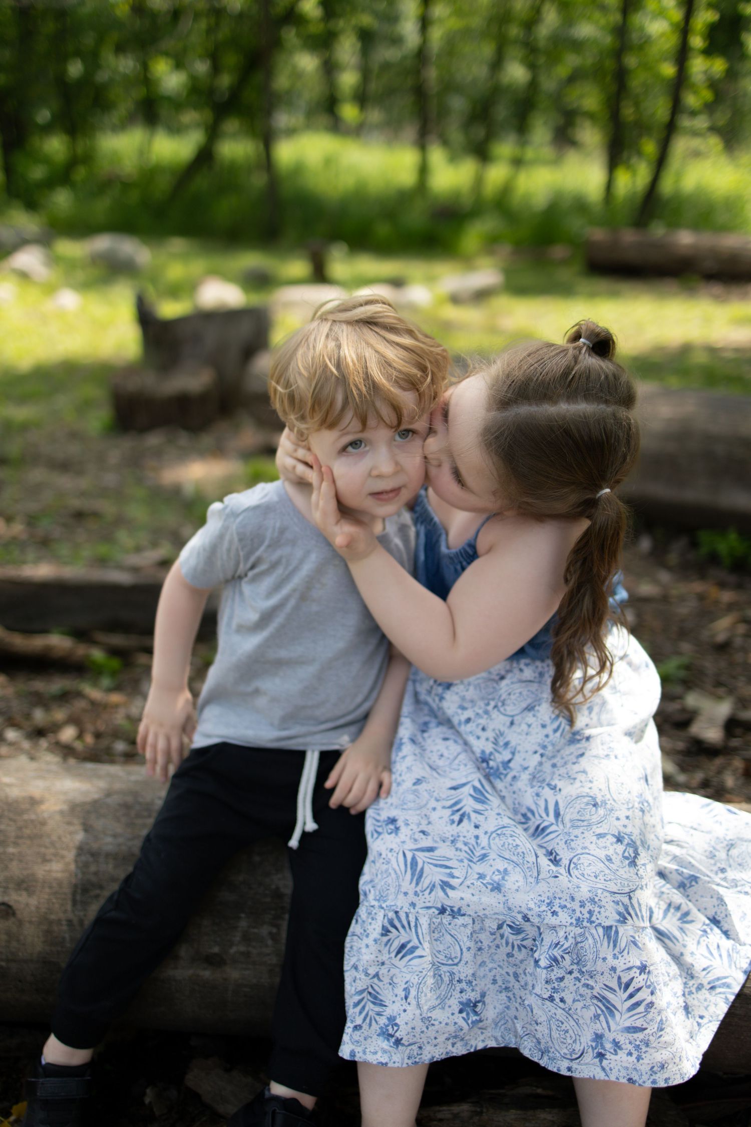 Two young children in grey and blue outfits sitting together on wooden logs in a sunny wooded outdoor setting.