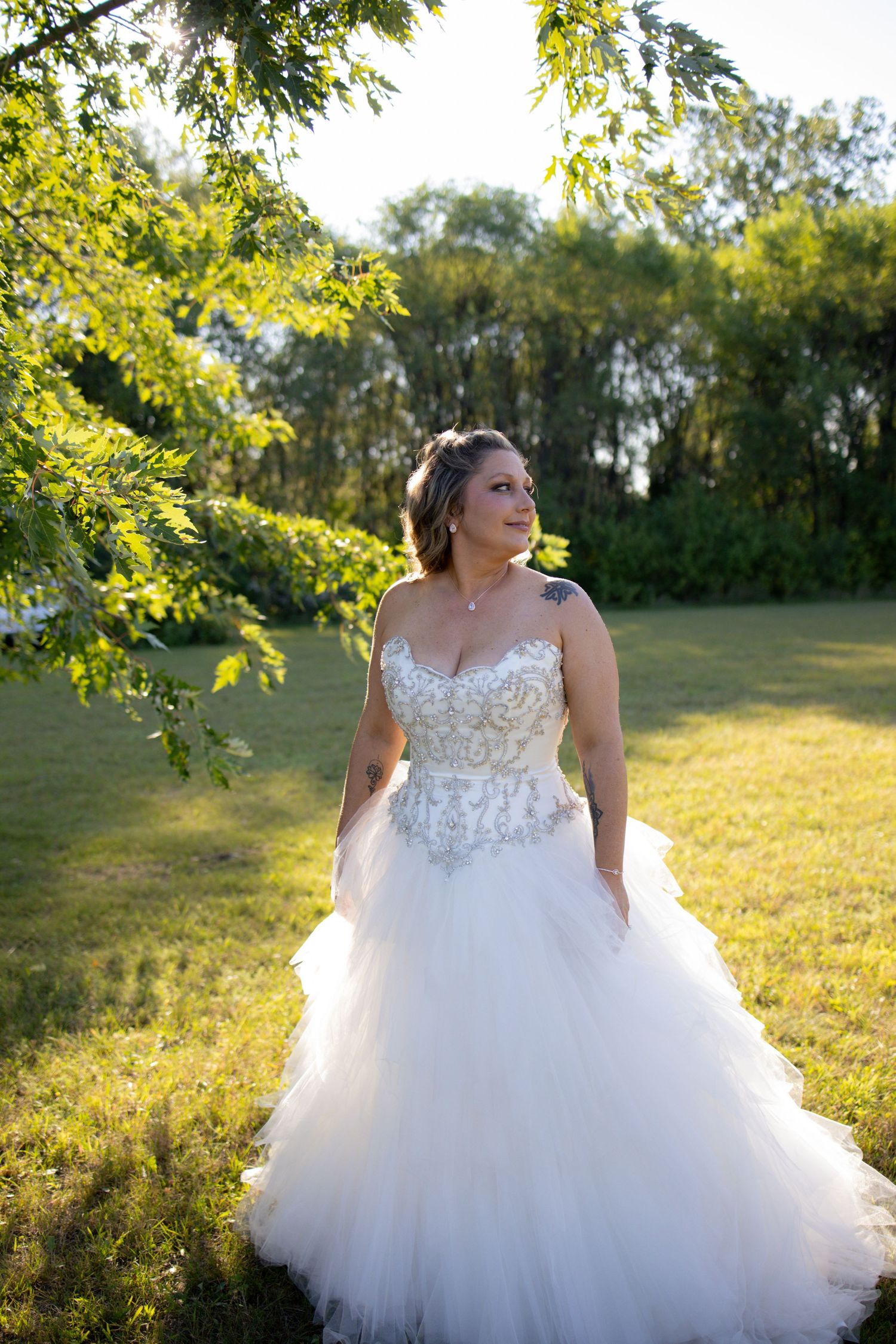 A bride in a stunning white ballgown with lace bodice poses in a sunlit field with golden light filtering through trees.