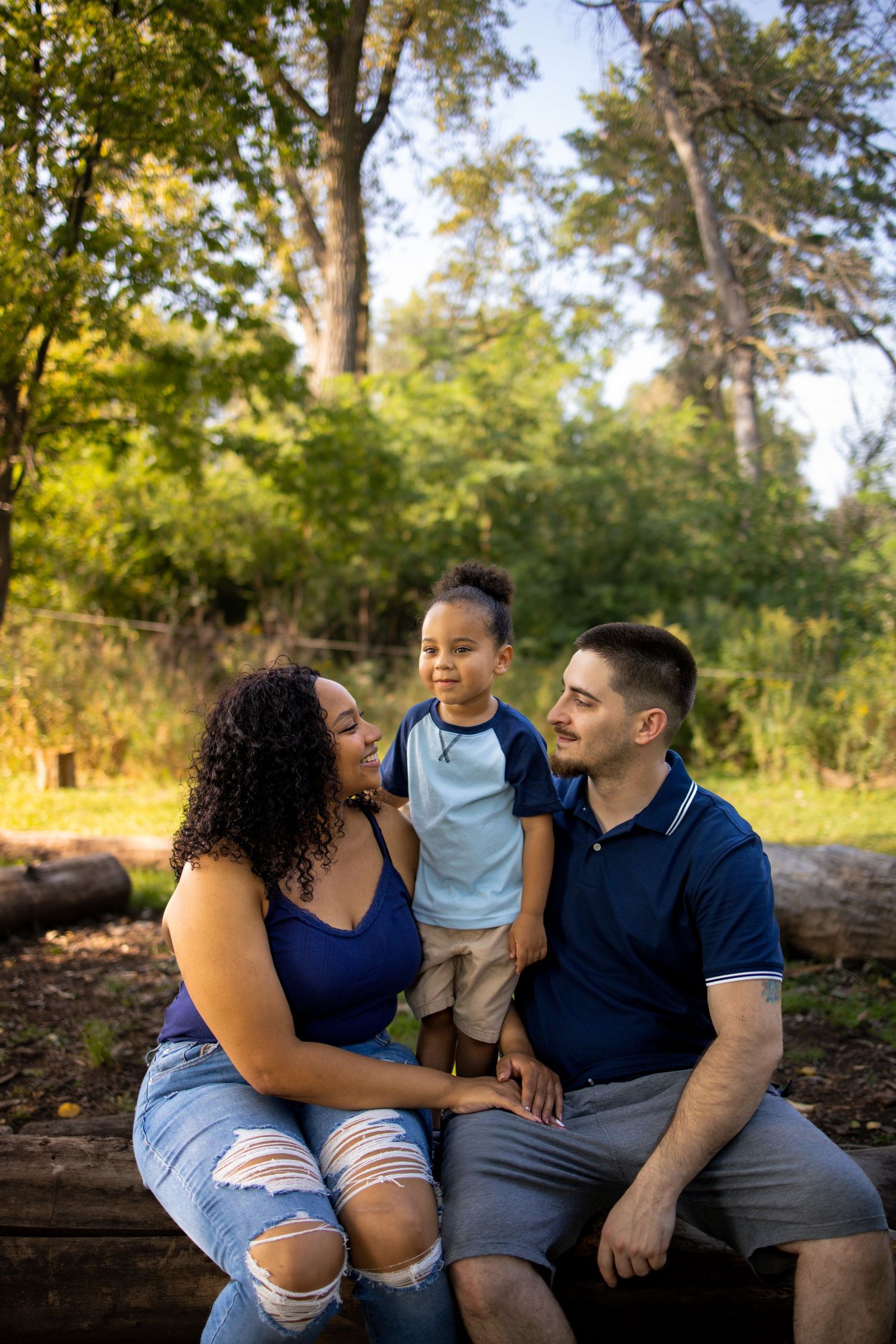 A loving family sits together outdoors on a wooden bench surrounded by lush green trees in warm sunlight.