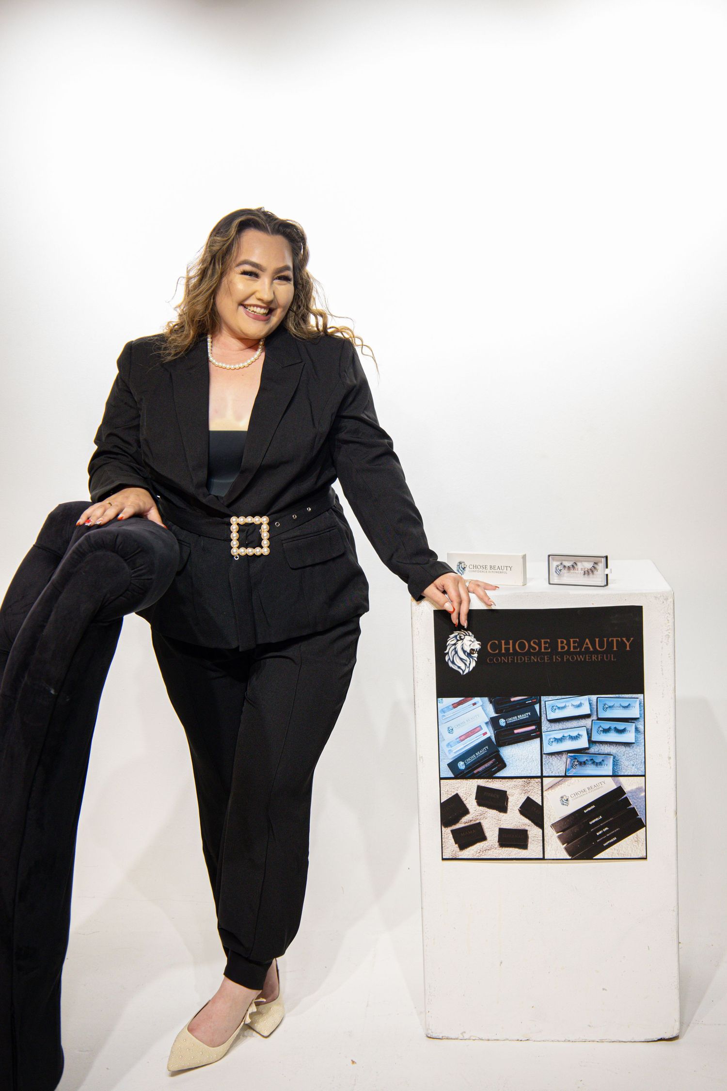 Professional in a black business suit poses next to promotional display featuring beauty product samples and marketing materials.