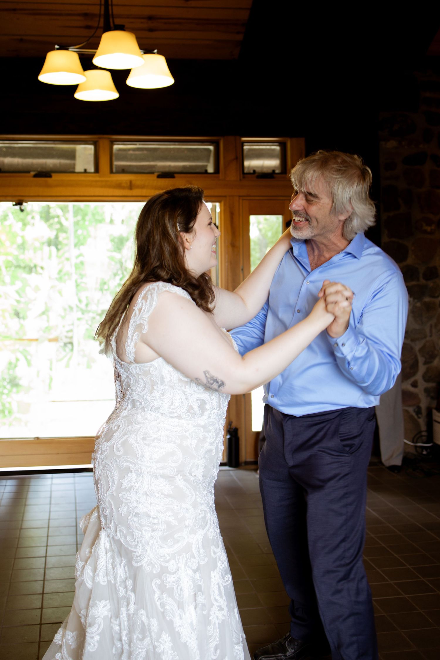 A bride in a white lace dress dances with someone in a blue shirt at an indoor reception venue with warm lighting.