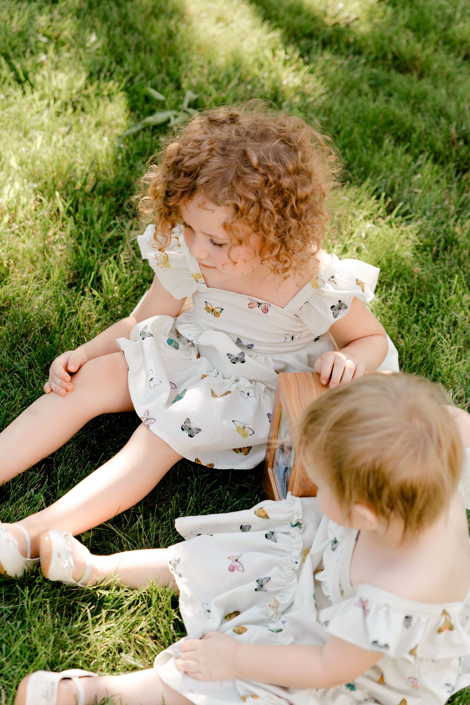Two small children in matching butterfly print dresses sit together on sunny grass.