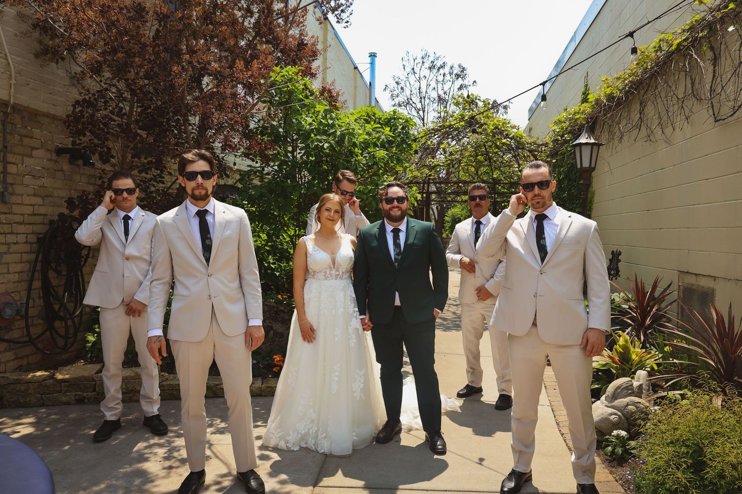 Wedding party wearing classic white suits and gowns poses together in a courtyard garden setting.