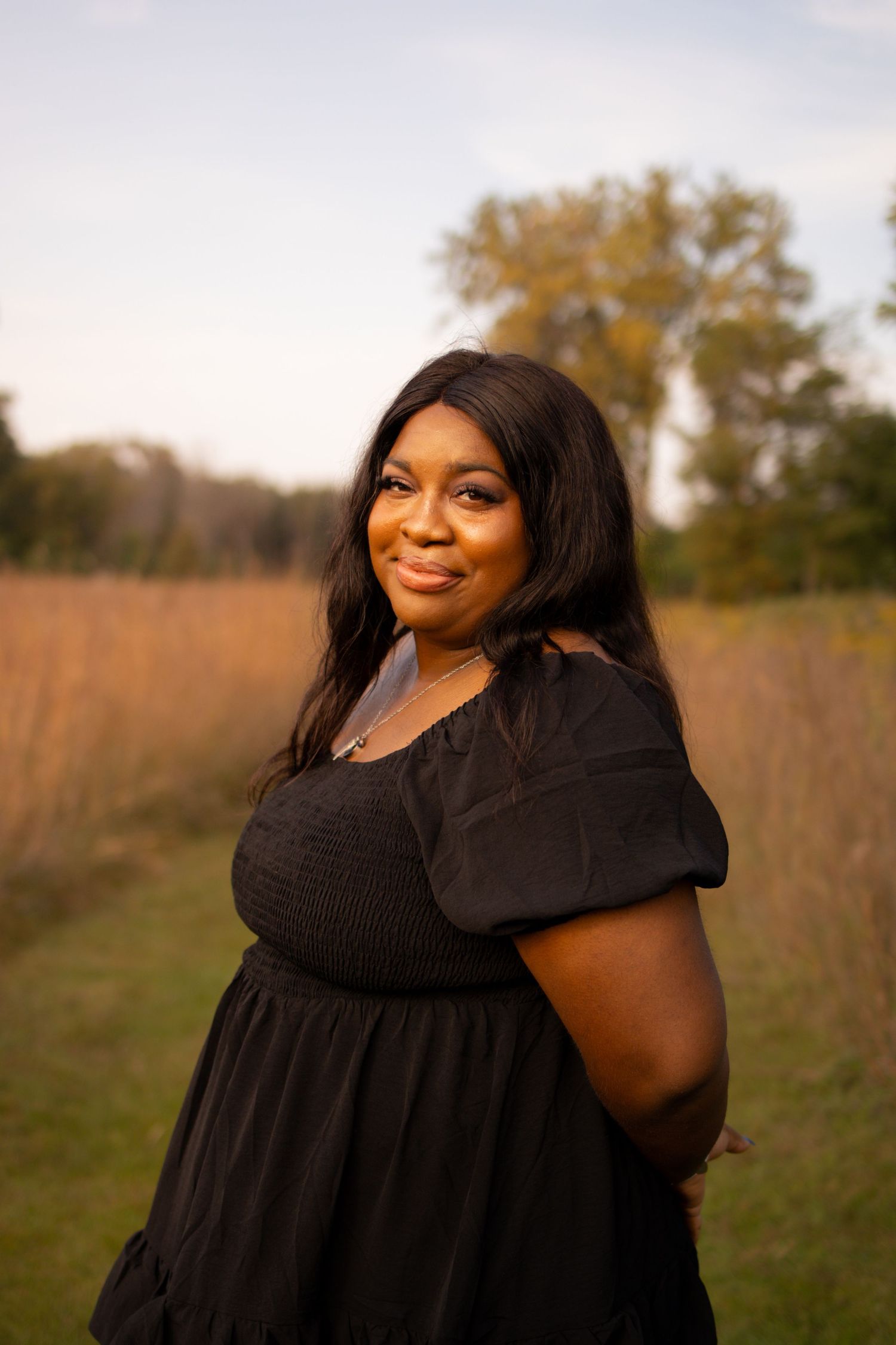 A woman in a black dress poses confidently in a golden field during sunset while smiling at the camera.