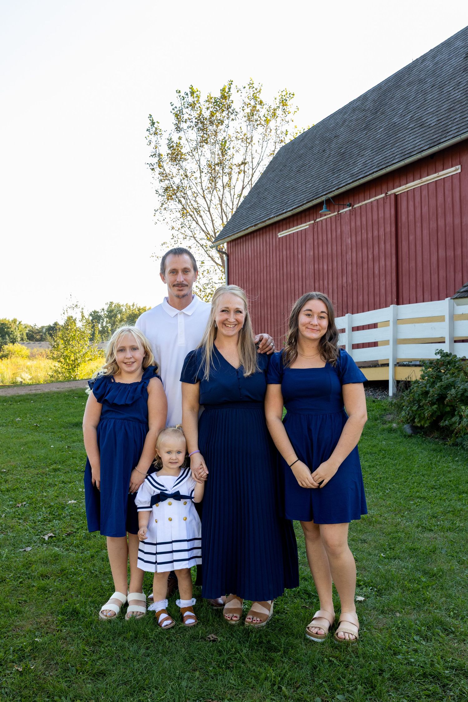 A family wearing coordinating navy blue and white outfits poses together on grass in front of a red barn.