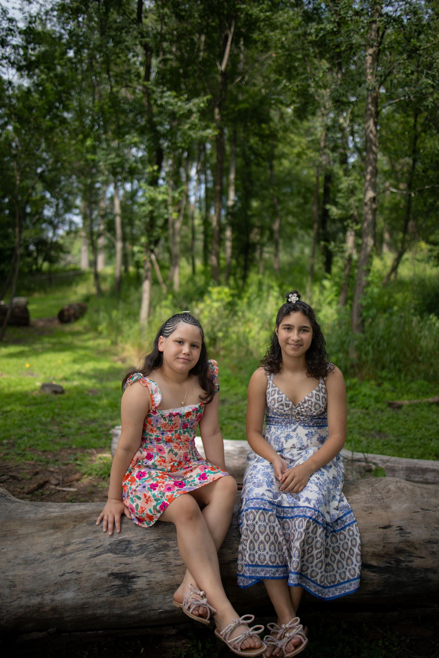 Two people in summer dresses sit together on a large rock in a wooded area on a sunny day.