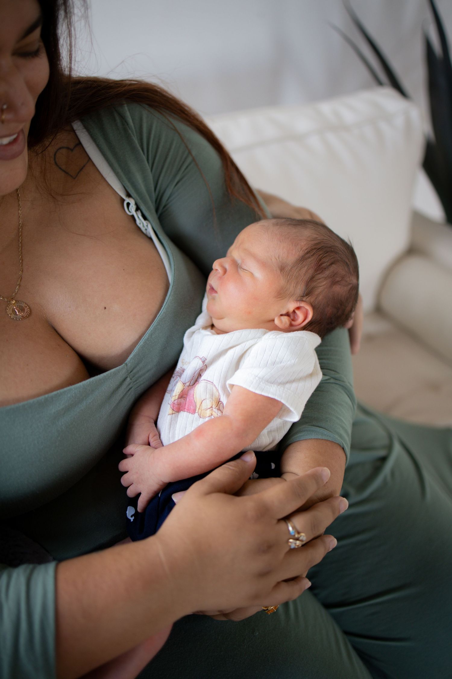 Sleeping newborn baby in white onesie rests peacefully against caregiver wearing green shirt.