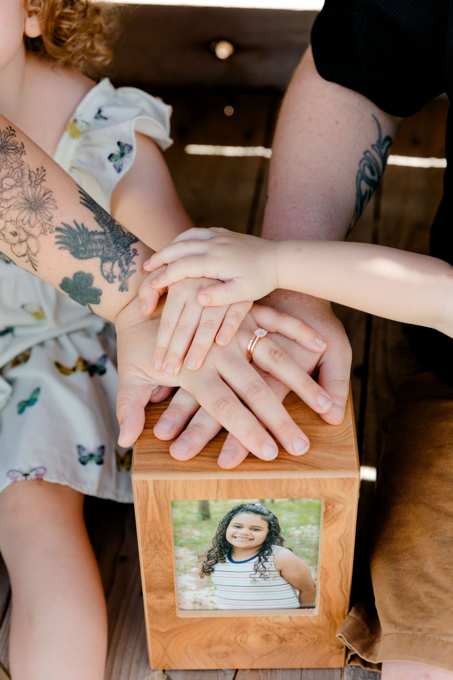 Close up of hands resting on wooden box with framed photo, showing tattoos and rings.