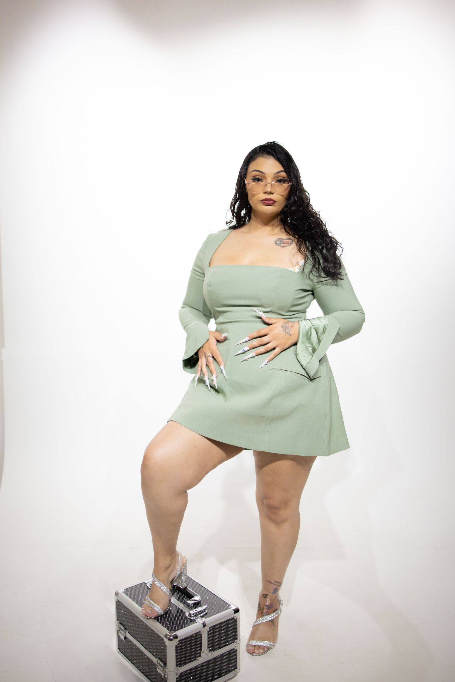 Model wearing a mint green mini dress with long bell sleeves poses on a storage case against white background.