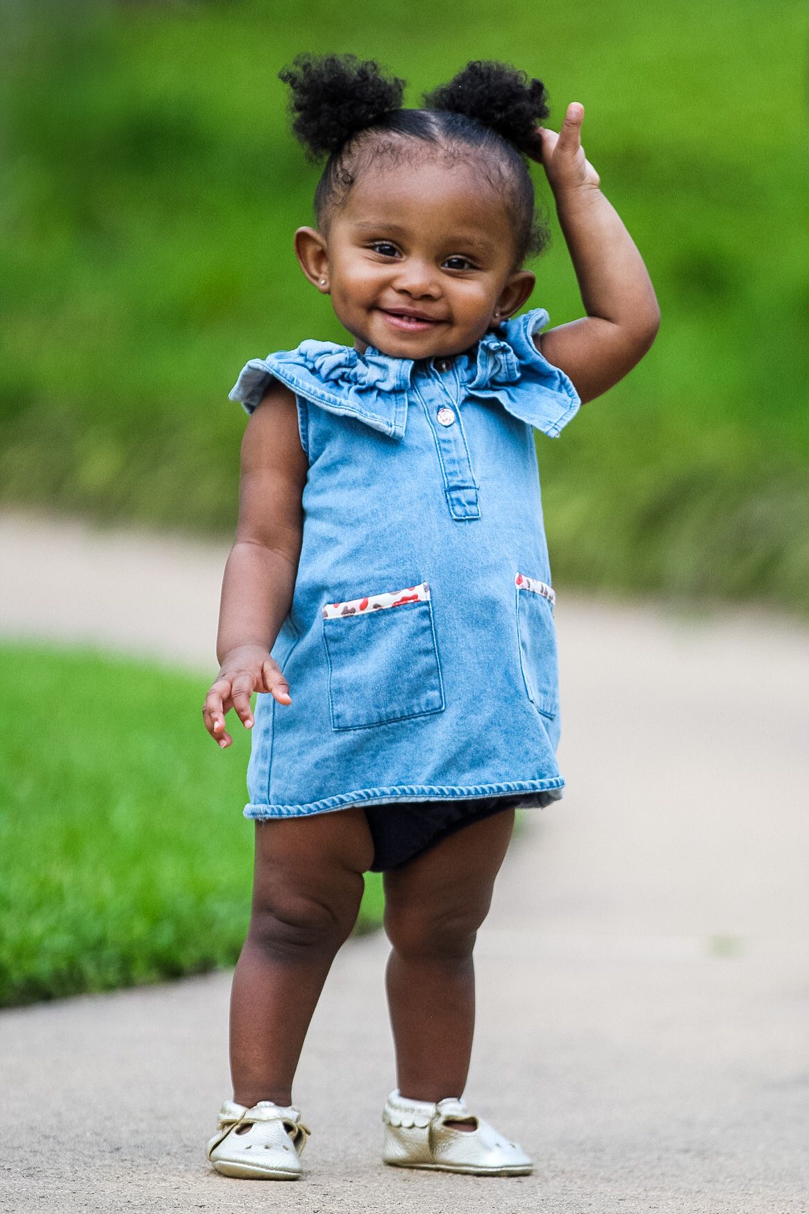 Toddler wearing blue denim dress and white shoes smiles while standing on a path with green grass background.