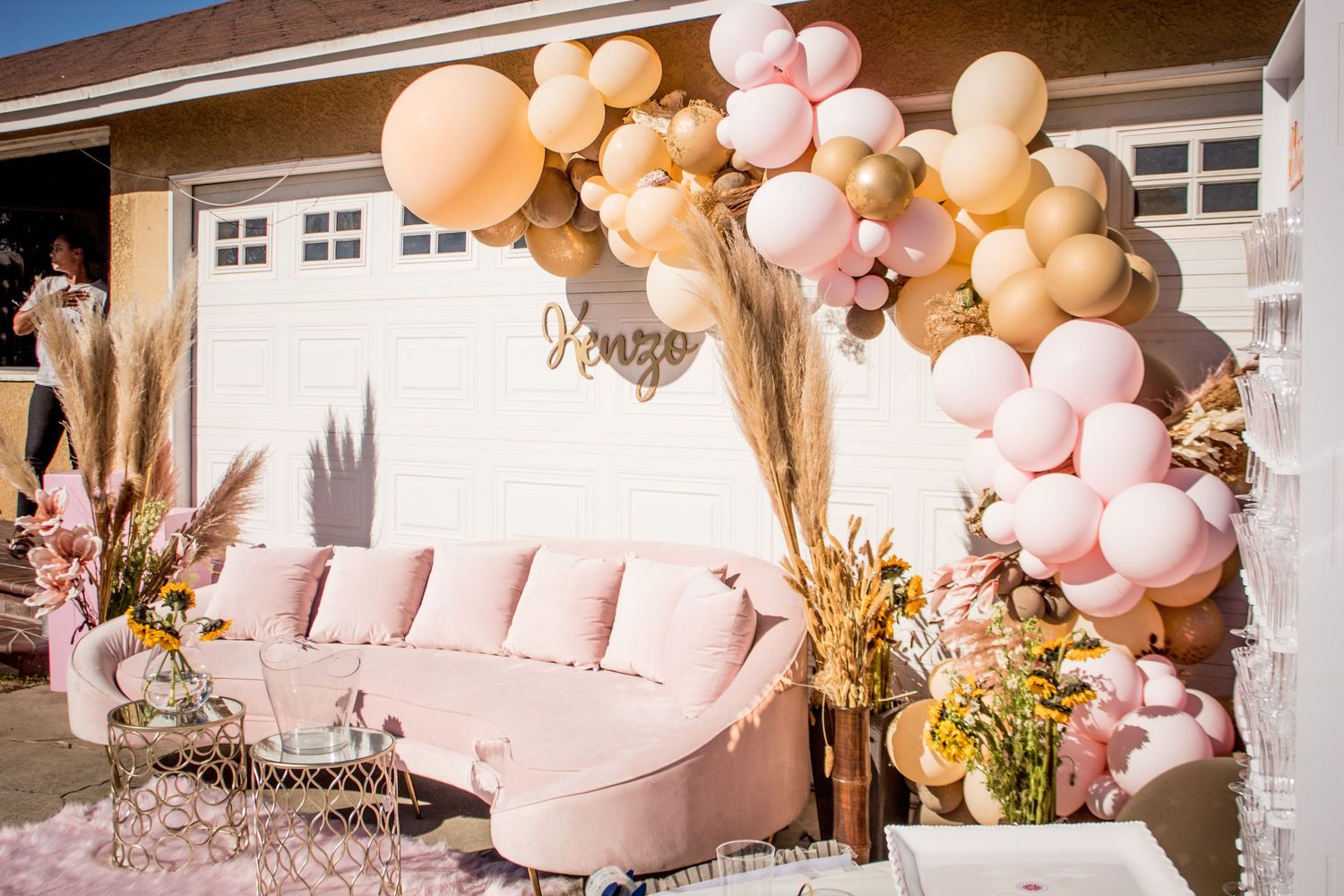 Pink and peach balloon arch decorates an outdoor party setup with a pink couch and dried pampas grass.