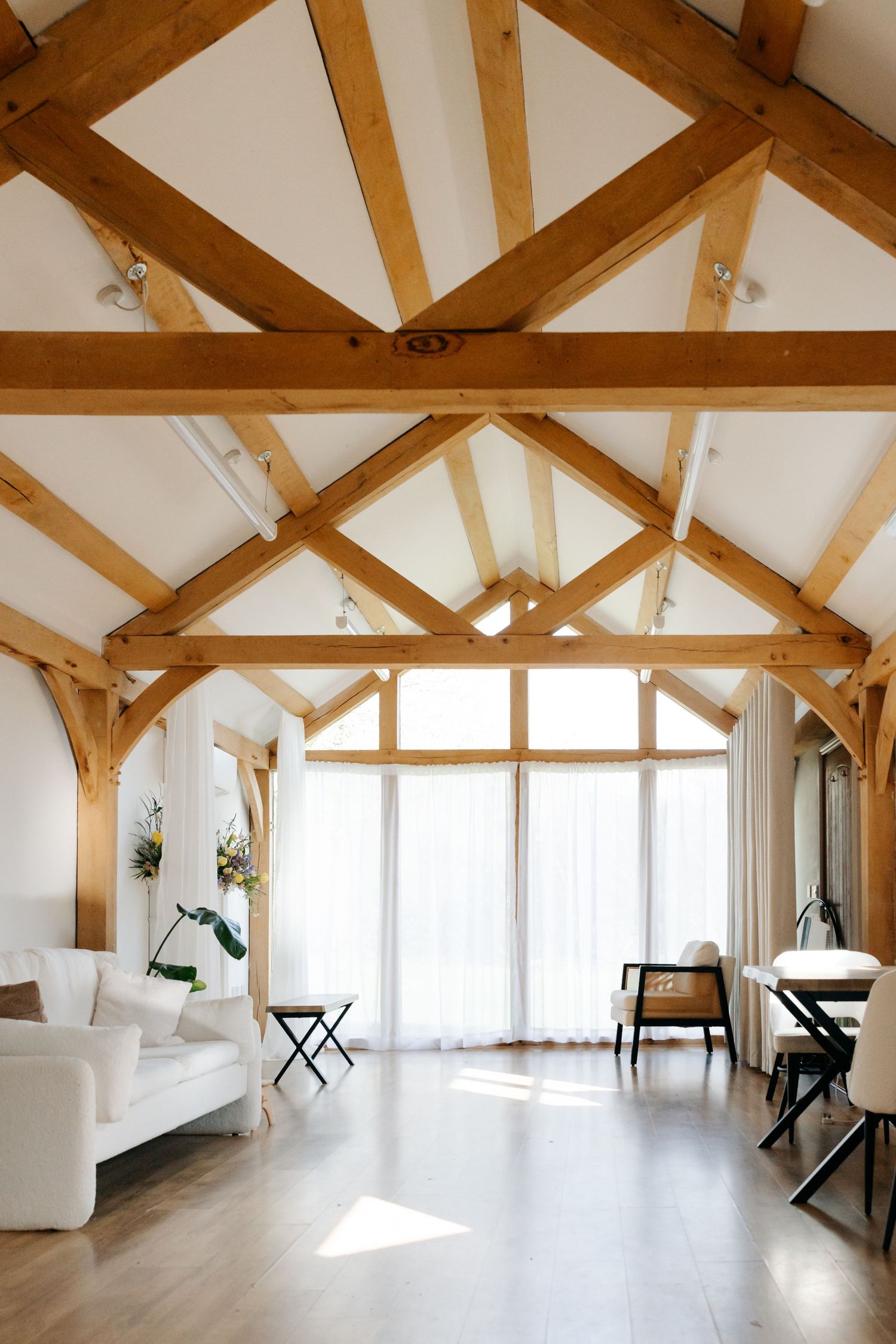 Bright barn conversion living room with exposed wooden roof trusses, white sofa, and large floor-to-ceiling windows.