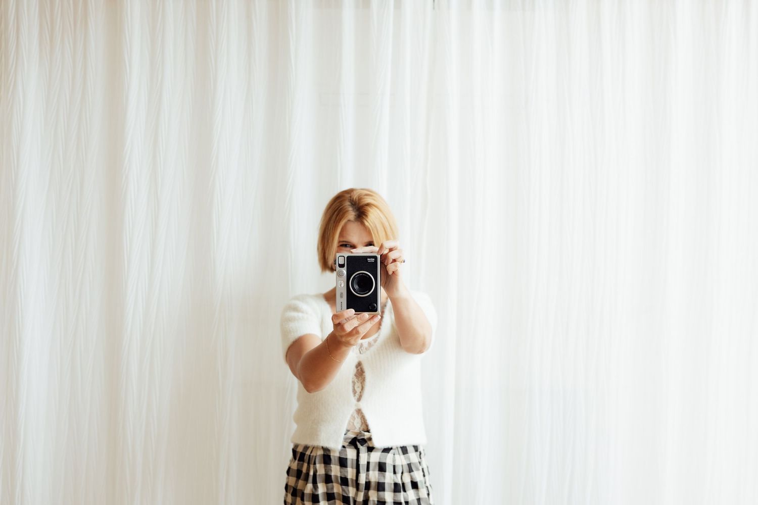 Woman in white top and plaid skirt holds vintage camera up to her face in front of white curtains.