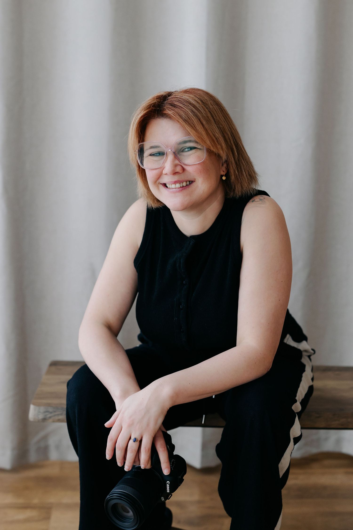 Smiling woman with glasses and red hair sits on bench holding a camera, wearing black outfit against neutral backdrop.