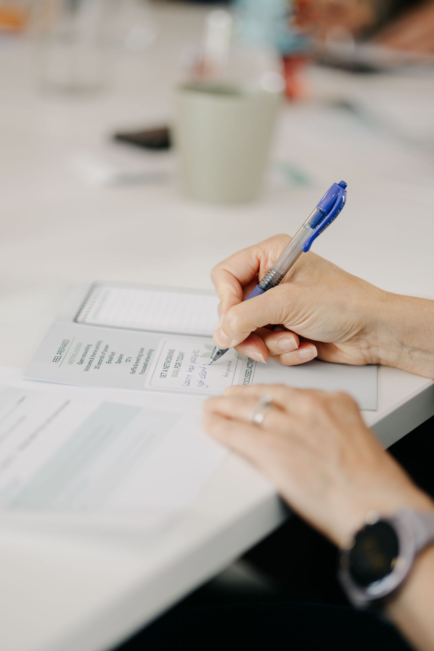 A hand holding a blue pen fills out a printed form on a white desk during a meeting.