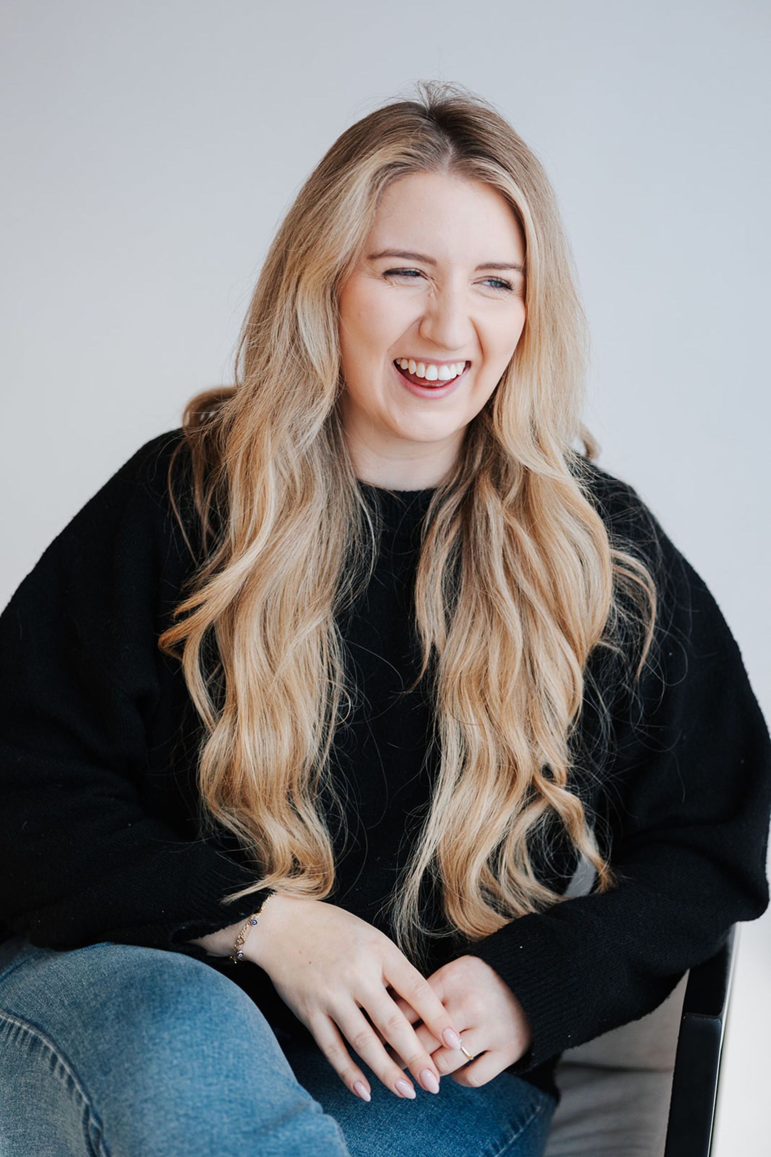 Smiling young woman with long blonde wavy hair wearing a black sweater and jeans, seated against a white background.
