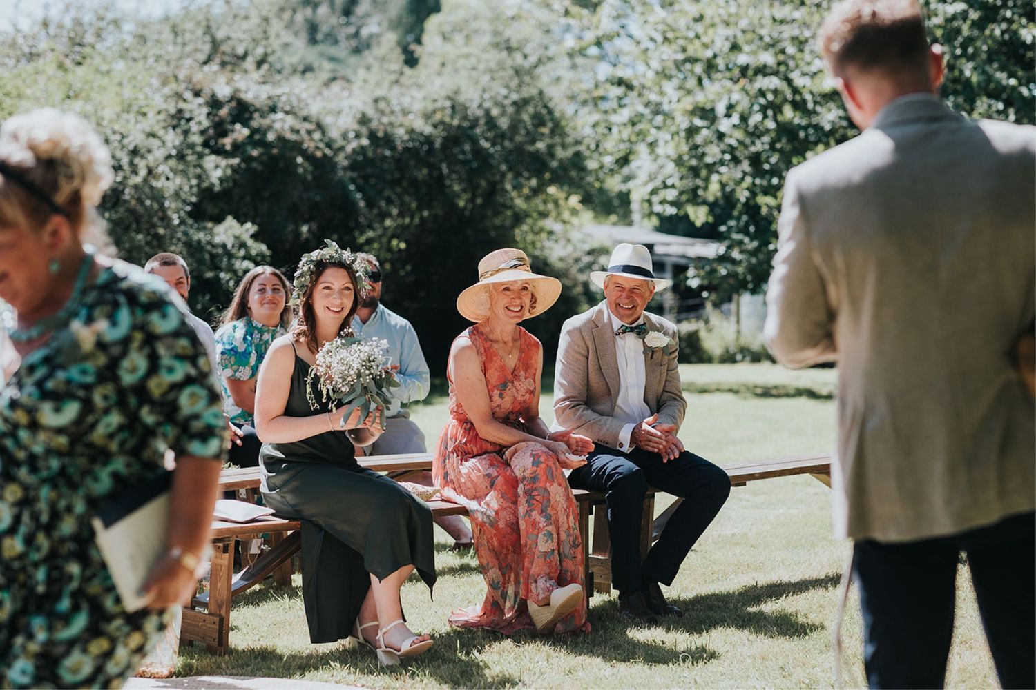 Wedding guests enjoy an outdoor ceremony on a sunny day in a garden setting.