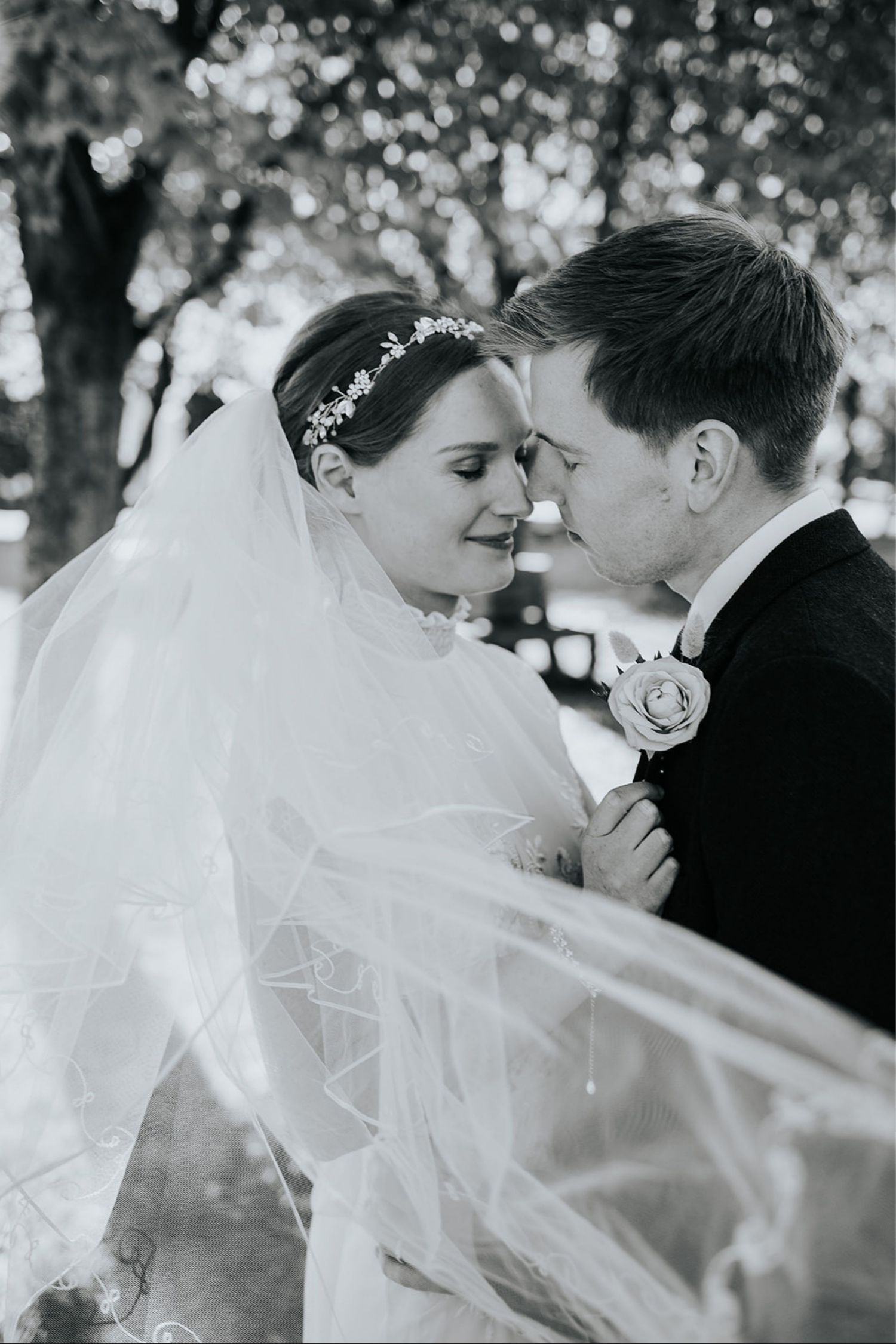 A tender black and white wedding moment capturing a couple in an intimate pose with flowing veil and floral headpiece outdoors.