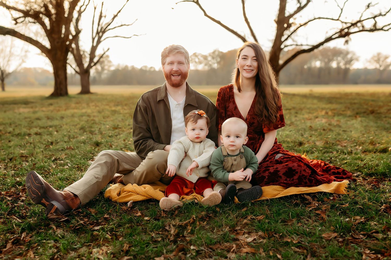 Family of four sitting on a blanket in a sunlit park, parents with two young children in autumn attire.