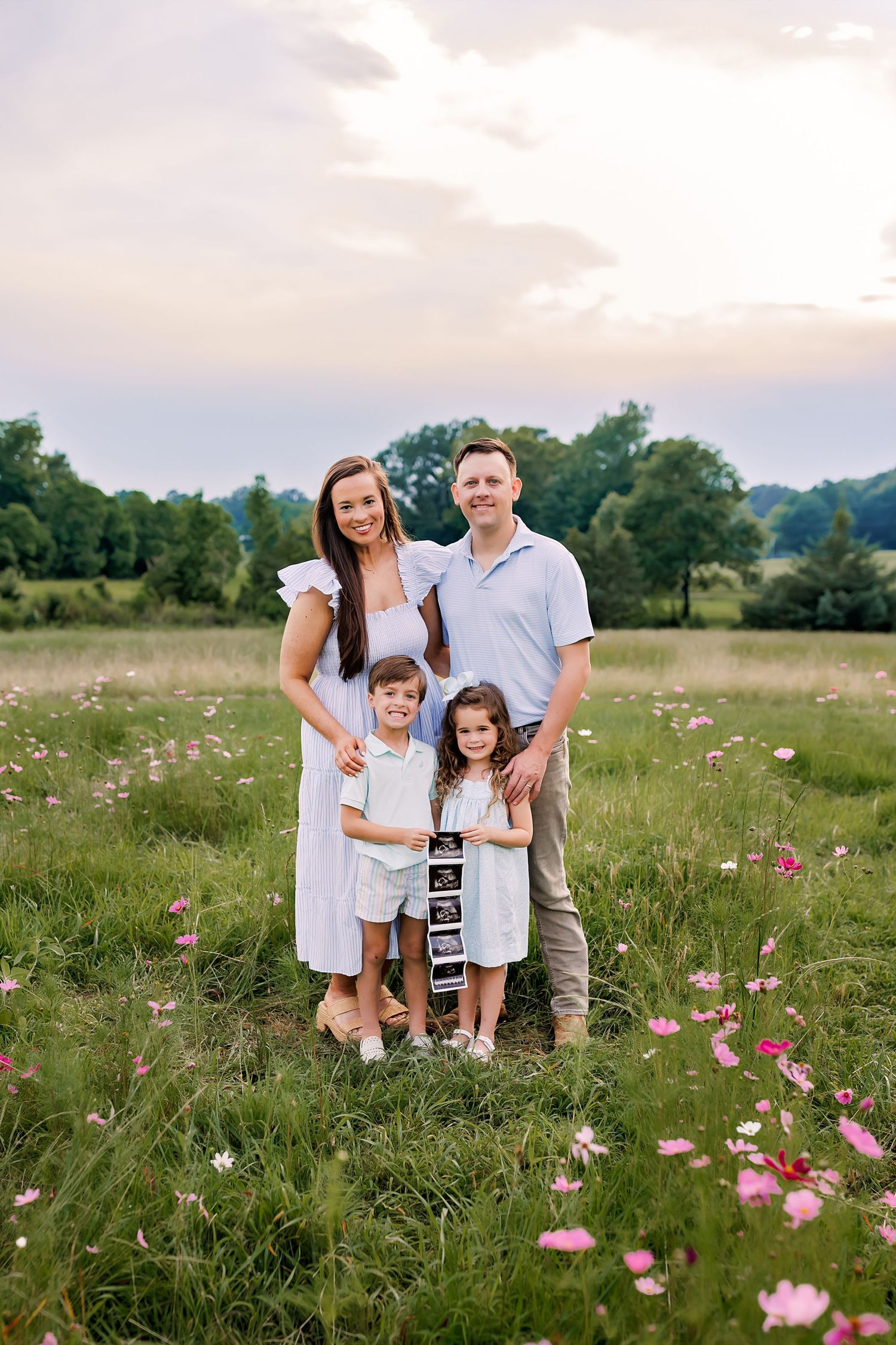 Happy family of four poses in blooming wildflower meadow holding ultrasound photo at sunset.
