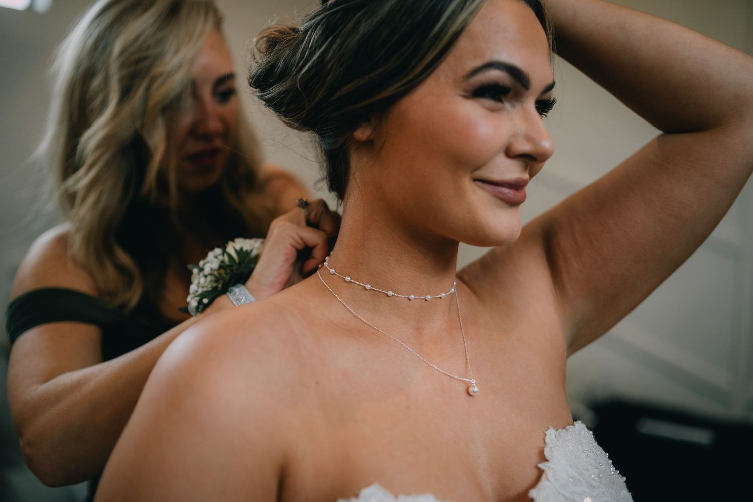 Bride smiling as bridesmaid fastens delicate pearl necklace, wearing strapless wedding gown with elegant updo hairstyle.
