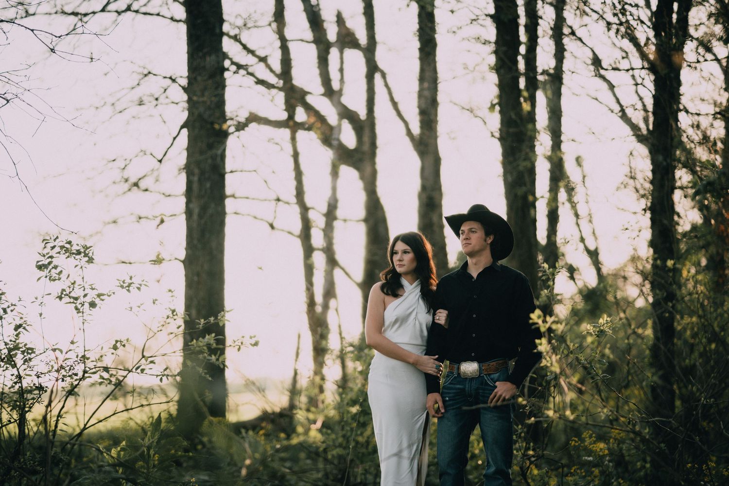 A romantic outdoor engagement photo shoot in a wooded area at sunset.