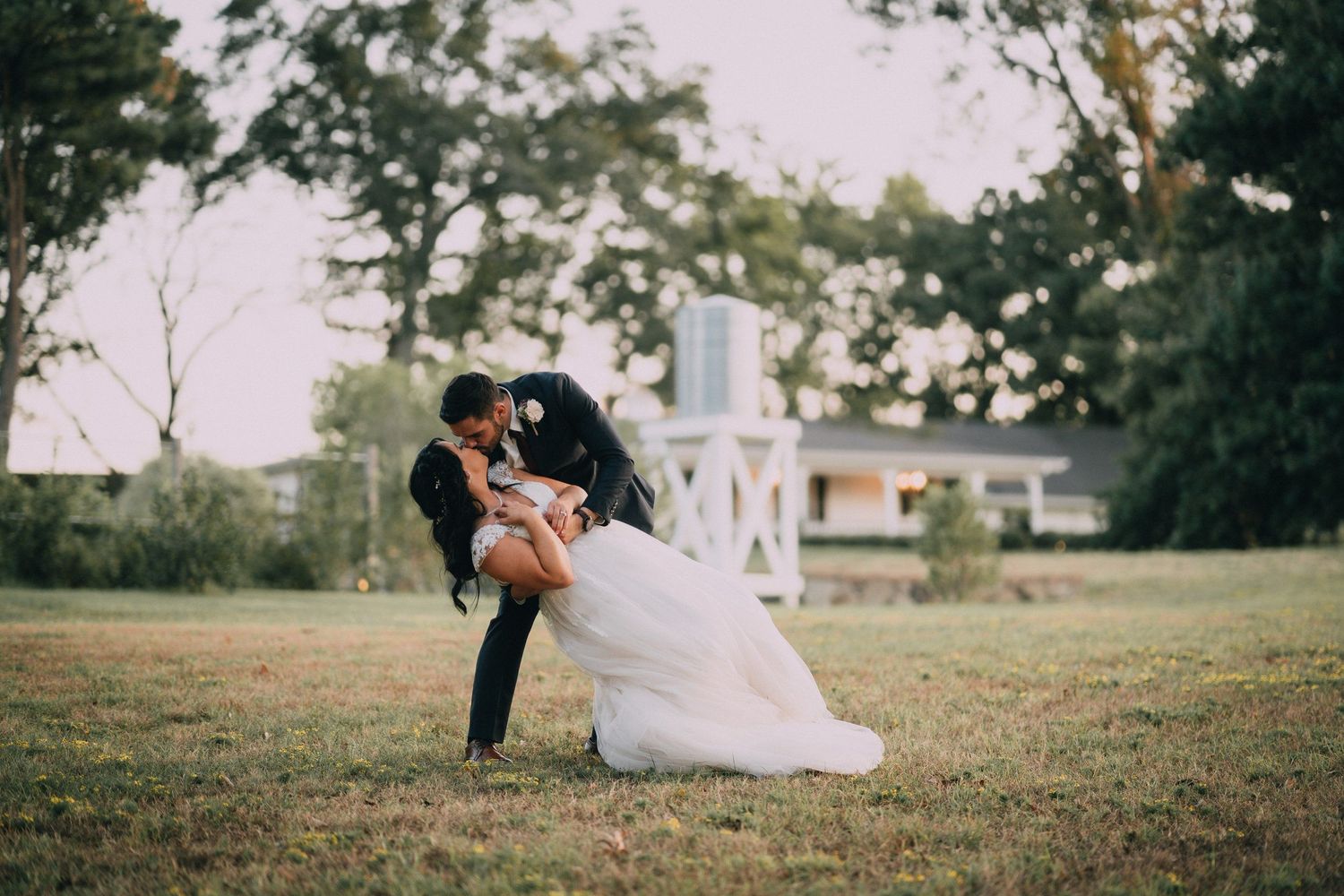 Romantic outdoor wedding scene with couple on grassy hill near white chapel.
