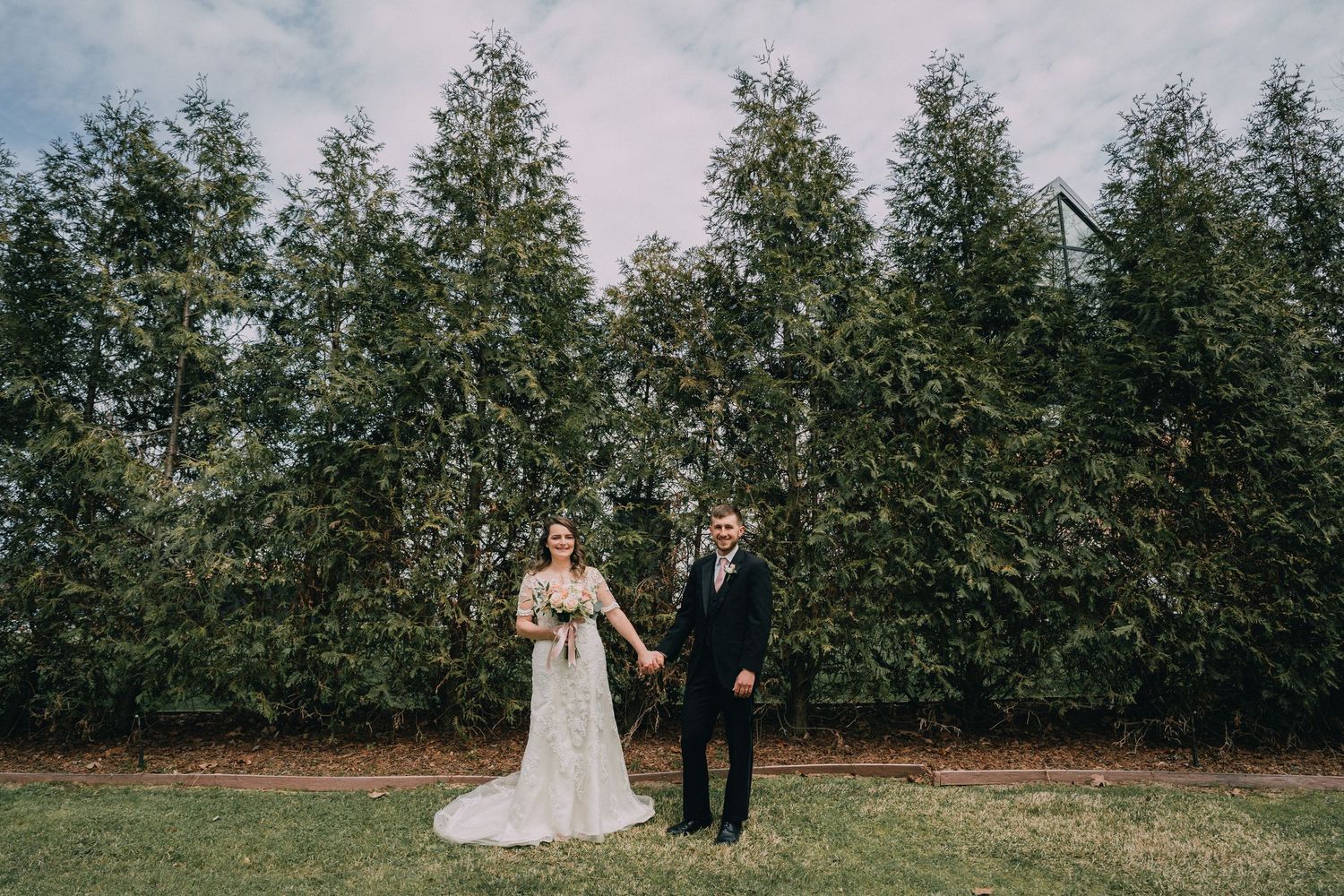 A wedding couple stands in front of a row of tall evergreen trees.