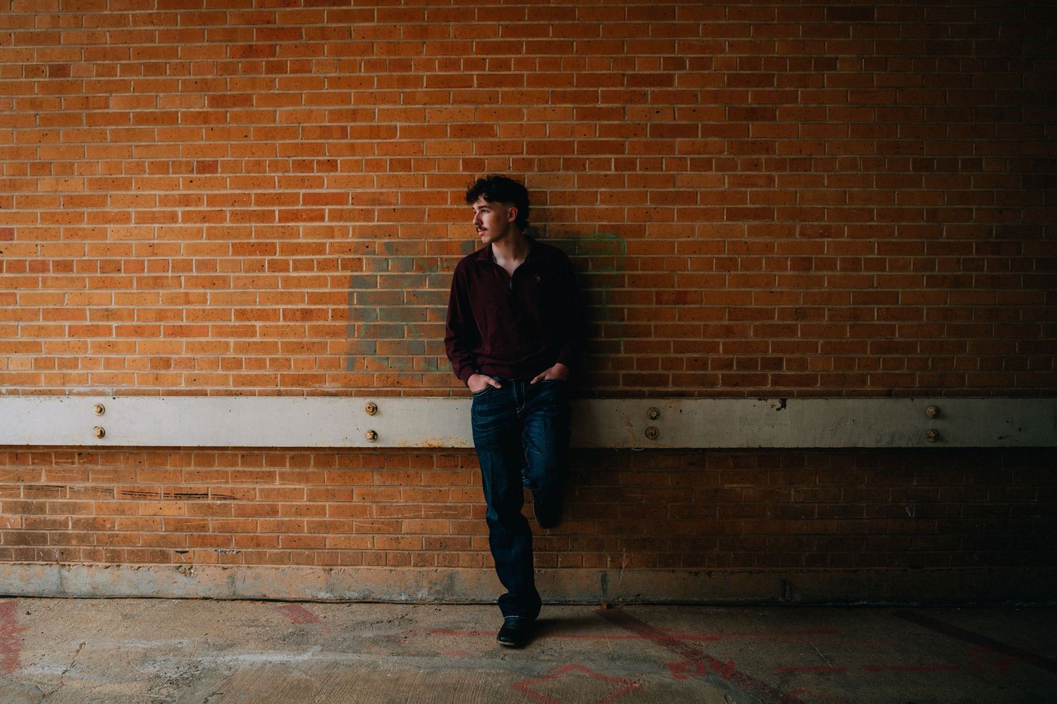 Young man in dark hoodie and jeans leaning against a brick wall outdoors.