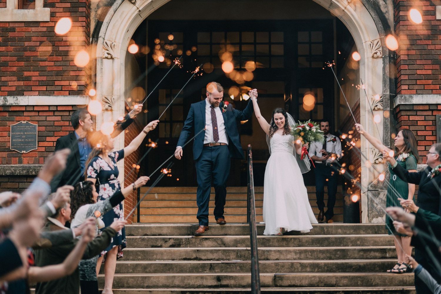 Wedding party exits brick church building under string lights at dusk.