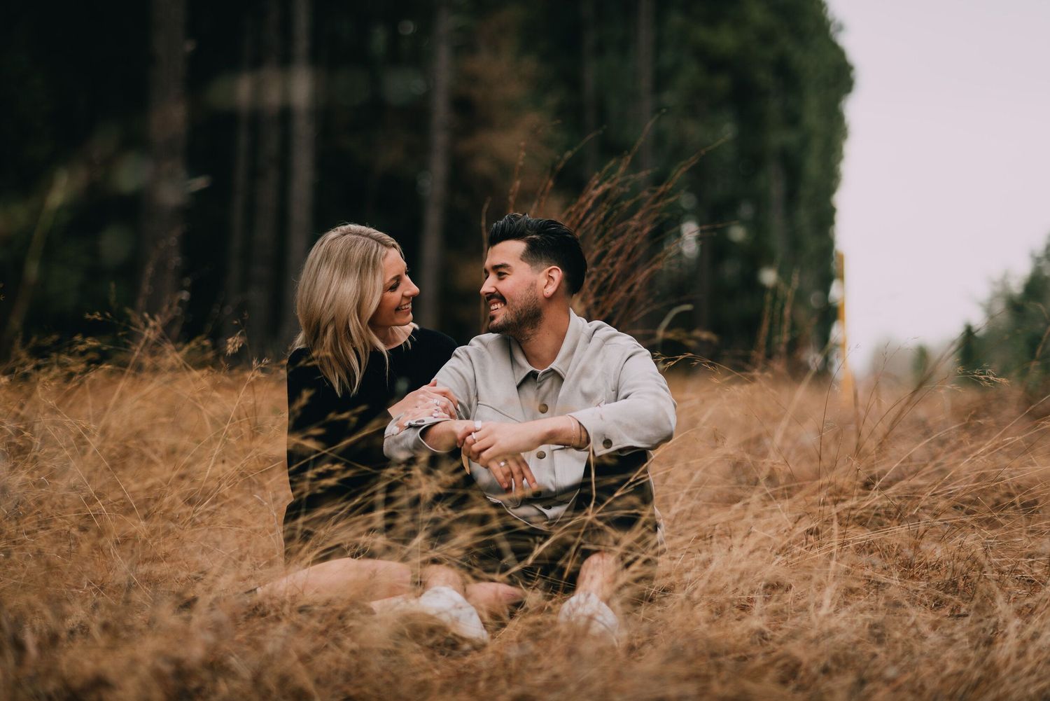 Couple embraces in a field of tall golden grass during sunset with trees in the background.