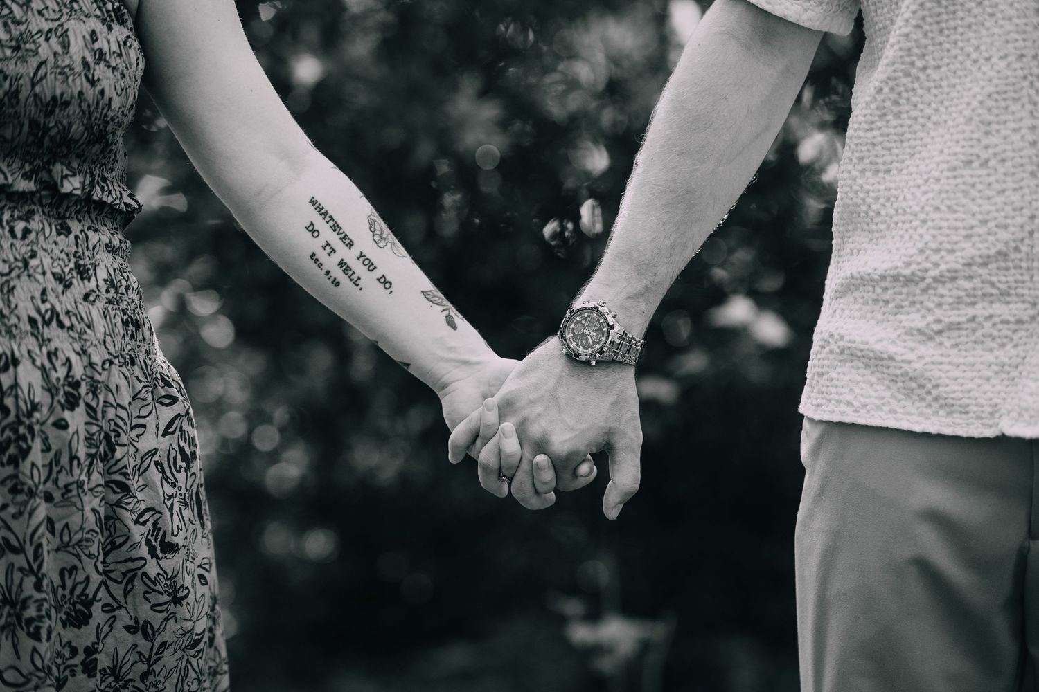 A black and white close-up of two people holding hands, showing a tattoo.