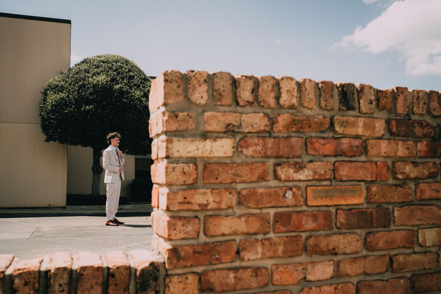 Man in light suit standing near rustic brick wall with green shrub and blue sky in background.