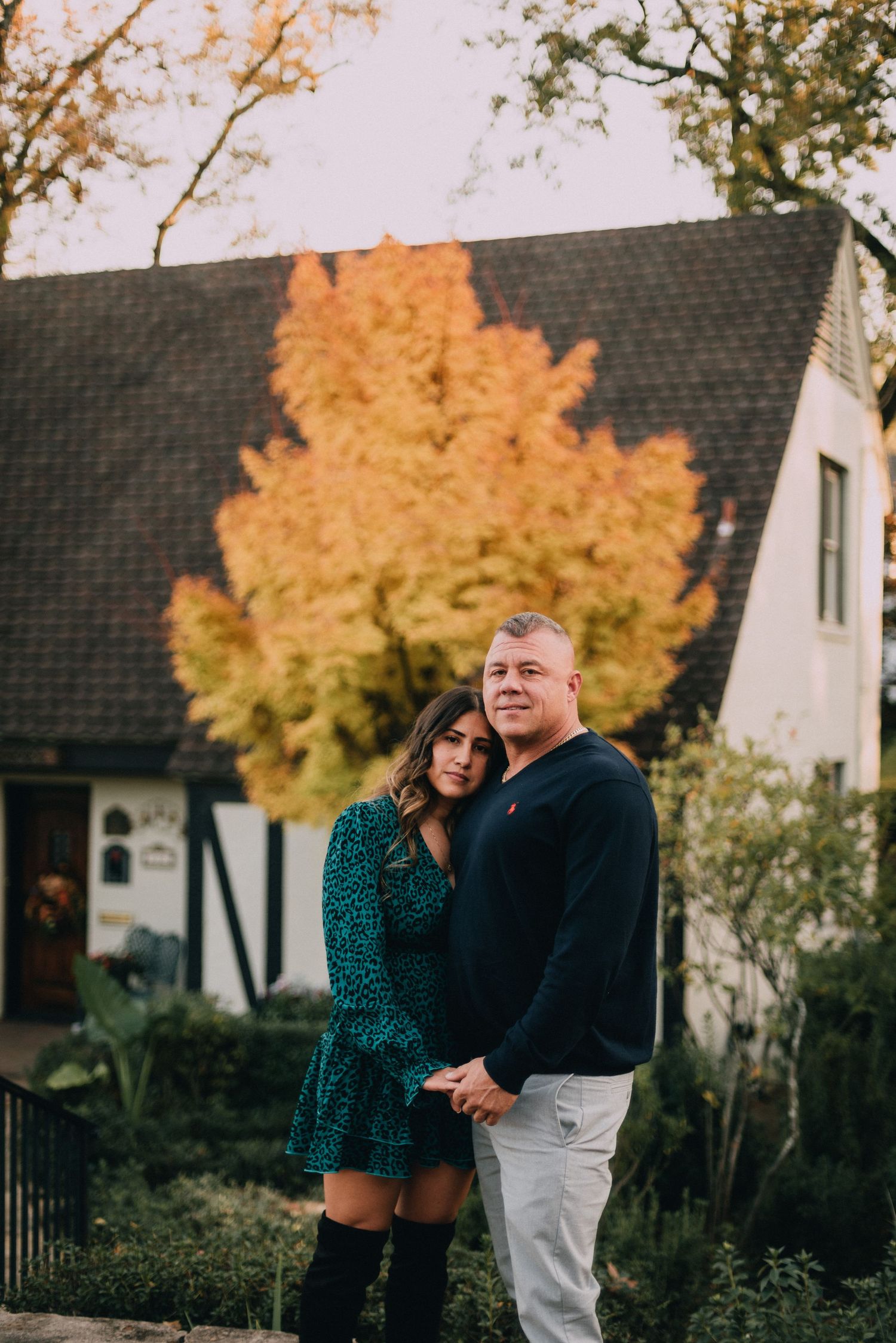 A couple shares an intimate moment in front of a house with autumn trees displaying vibrant orange foliage.