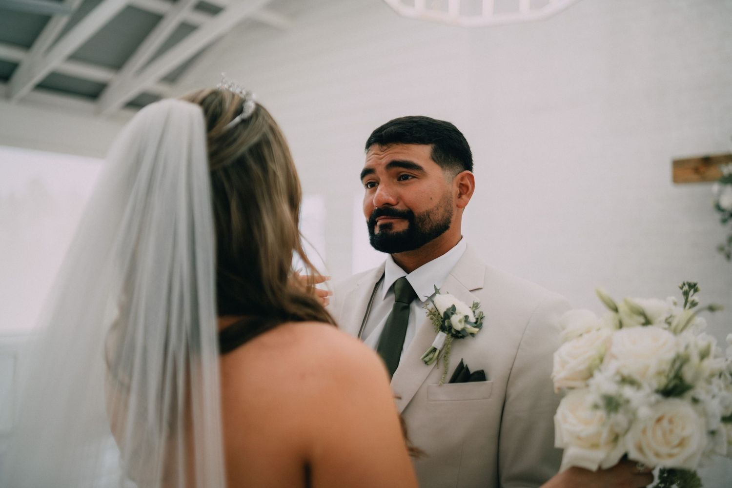 Groom in beige suit with green tie smiling at bride in white veil during wedding ceremony.
