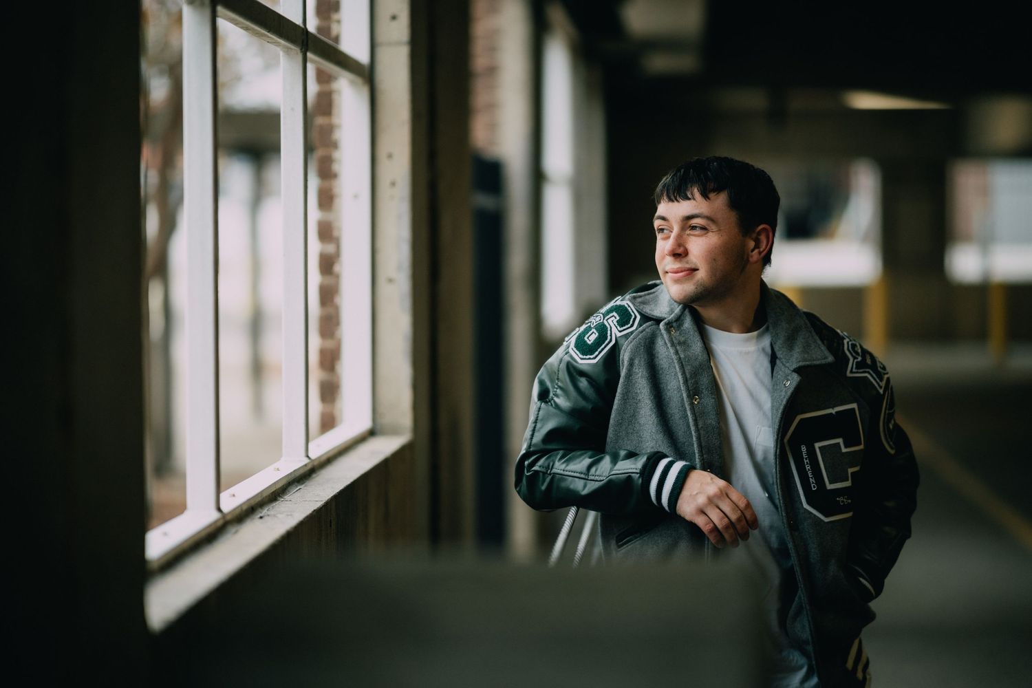 Young man in a varsity jacket leaning against a window in an urban building, gazing thoughtfully outside.