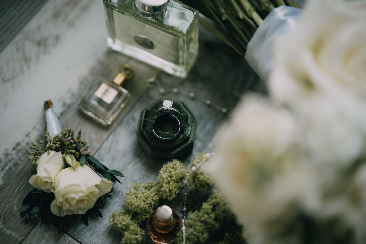 Elegant wedding flat lay with perfume bottles, white roses, a ring, and boutonniere on a rustic wooden surface.