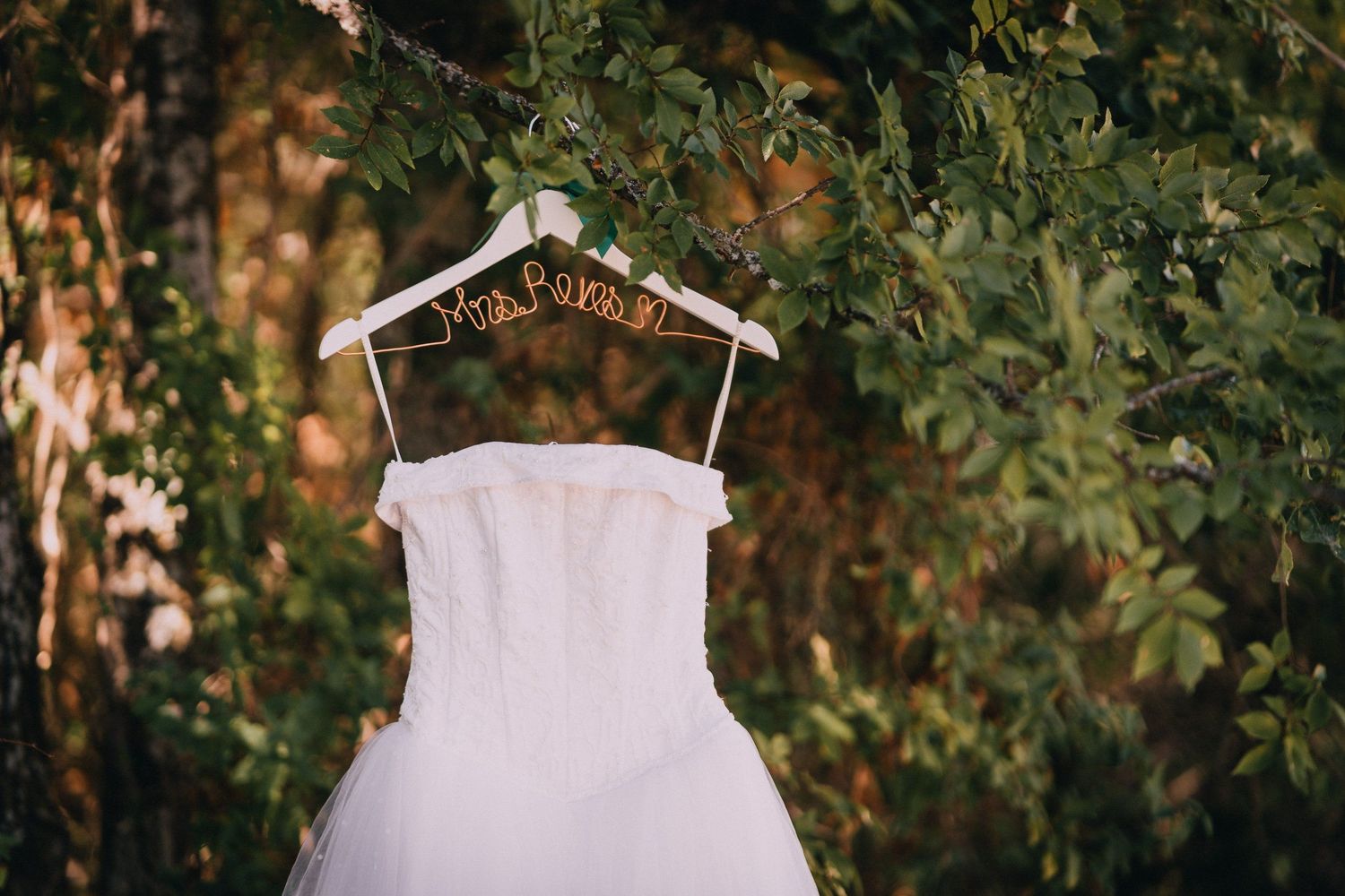 White wedding dress hanging on wooden hanger against green foliage background.