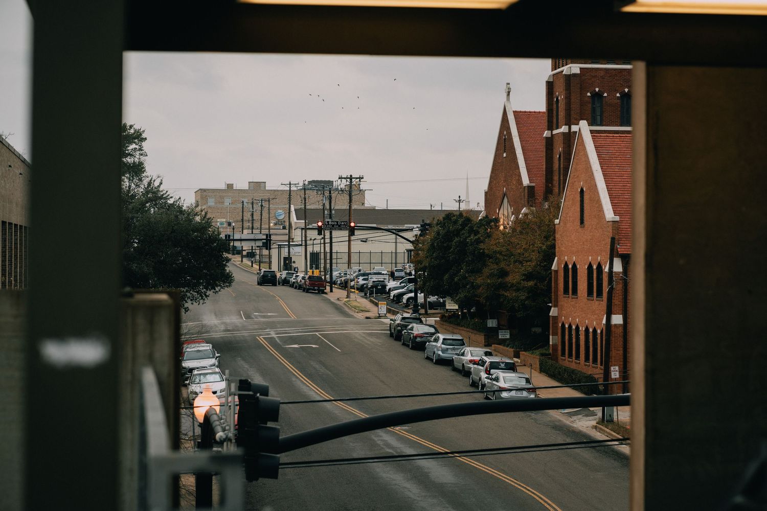 Urban street view framed by a window, featuring a red brick church, parked cars, and city buildings on a cloudy day.