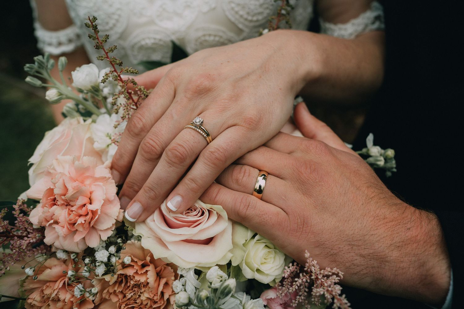 Wedding rings and hands resting on white lace dress with pink floral bouquet.