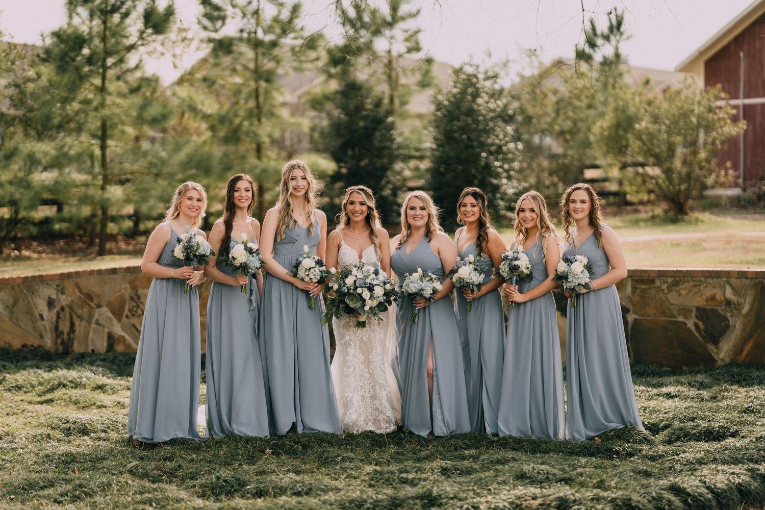 Row of bridesmaids in light blue dresses pose together on grassy lawn near rustic barn.