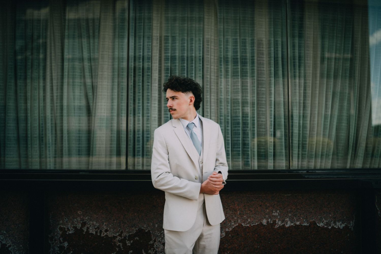 A man in a light beige suit and blue tie stands confidently outdoors near a modern glass building.