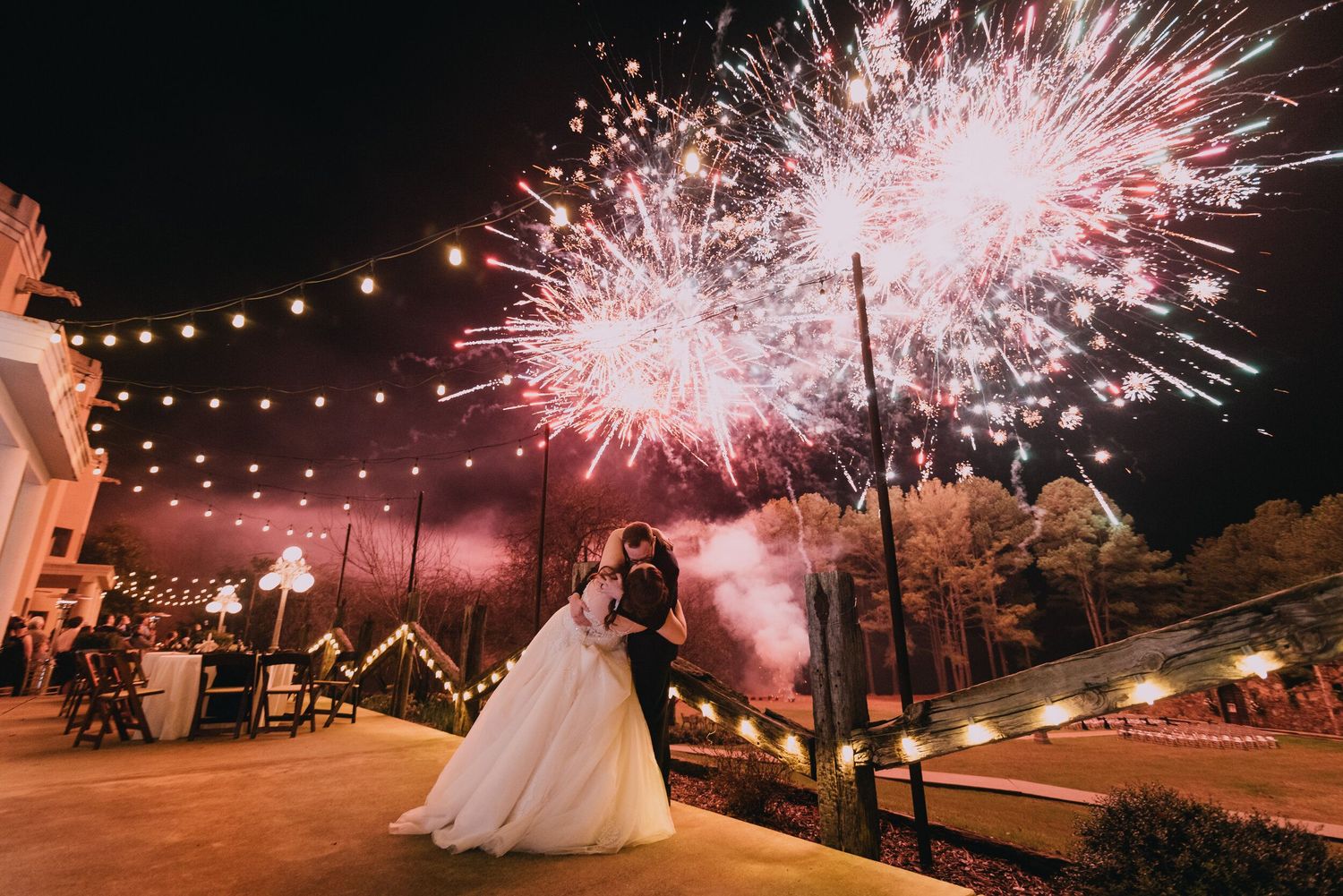 Pink and white fireworks display illuminating night sky at outdoor wedding reception.